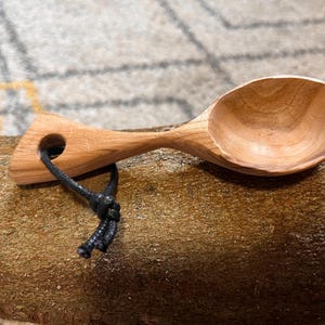 Könnte beinhalten: Ein kleiner Holzlöffel mit einem schwarzen Schnurstiel. Der Löffel ist aus hellem Holz und hat eine abgerundete Schale.