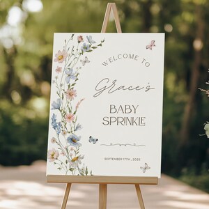 May include: A cream-colored sign on a wooden easel, with the words "Welcome to Grace's Baby Sprinkle" in elegant script. The sign is decorated with watercolor flowers and butterflies. The date "September 17th, 2025" is also displayed.