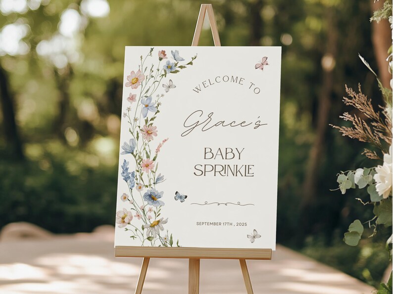 May include: A cream-colored sign on a wooden easel, with the words "Welcome to Grace's Baby Sprinkle" in elegant script. The sign is decorated with watercolor flowers and butterflies. The date "September 17th, 2025" is also displayed.