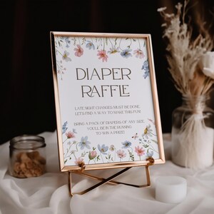 May include: A white sign with a gold frame and floral border. The sign reads "Diaper Raffle" and includes the text "Late night changes must be done. Let's find a way to make this fun! Bring a pack of diapers of any size. You'll be in the running to win a prize!"