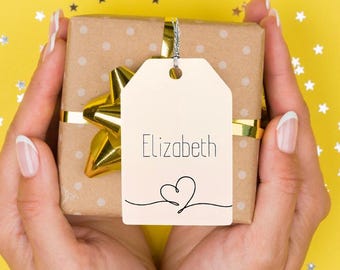 May include: A wrapped gift with a gold ribbon and bow, held in two hands. The gift is wrapped in brown paper with white polka dots. A tag with the name "Elizabeth" and a heart is attached.