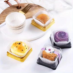 May include: Various baked mooncakes in clear plastic boxes with paper liners. The mooncakes are different colours and designs, some with sesame seeds. A stack of white paper baking cups is also visible. The presentation is on a white surface.
