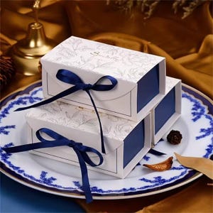 May include: Three white gift boxes with navy blue interiors and lids, stacked on a decorative plate. The boxes feature a floral pattern and are tied with navy blue ribbons. The plate has a blue and white floral design.