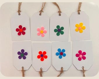 May include: Eight white gift tags with colorful flower embellishments. Each tag has a different colored flower: red, pink, green, yellow, purple, red, blue, and pink. The tags are strung with natural twine.