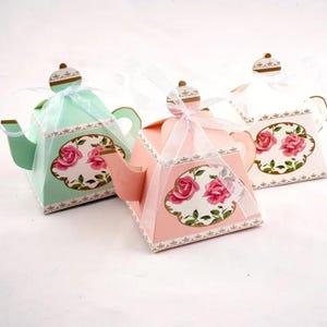 Op de afbeelding: Drie decoratieve theepotvormige dozen in pastelkleuren: mintgroen, roze en gebroken wit. Elke doos heeft een bloemenontwerp met roze rozen en een gouden rand. De dozen zijn vastgebonden met witte linten, geschikt voor cadeaus of feestartikelen.