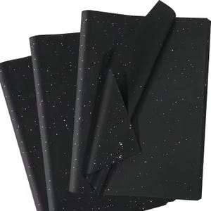 May include: Black tissue paper with a speckled glitter design. The sheets are folded and layered, showing the texture and sparkle. Suitable for gift wrapping or craft projects.
