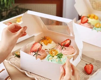 May include: White bakery box with a clear window, holding six cupcakes with pink, green, and white frosting. Each cupcake is topped with fresh fruit, including strawberries and orange slices. The box sits on a decorative tray.