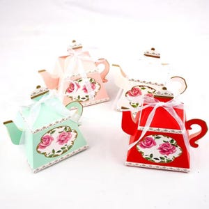 May include: Four decorative teapot-shaped gift boxes in pastel colours: pink, white, mint green, and red. Each box features a floral design with pink roses and a white ribbon. The teapots have gold accents.