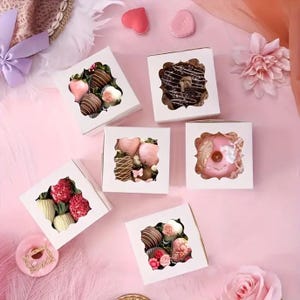 Op de afbeelding: Overzicht van witte dozen met heldere ramen, elk met chocolade-aardbeien en een donut. De aardbeien zijn gedoopt in verschillende chocolades en versierd. De achtergrond is zachtroze met decoratieve elementen.
