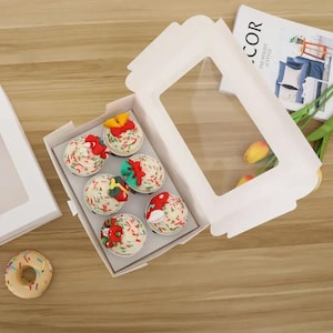 Puede incluir: Una caja de panadería blanca con una ventana transparente, que contiene seis cupcakes decorados con chispas de colores y decoraciones festivas. También se ve un solo donut y una revista.