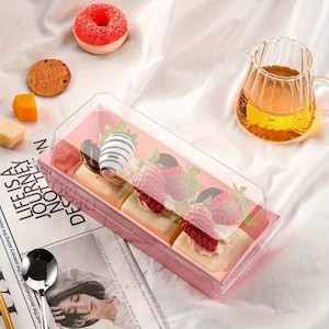 Może przedstawiać: Różowe prostokątne pudełko na ciasto z przezroczystą pokrywką zawiera małe ciasta z truskawkami i czekoladą. Pączek, ciastko i inne słodycze są rozrzucone. Widoczny jest również szklany dzbanek z herbatą i łyżka.