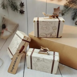 May include: Three decorative cardboard boxes designed as vintage suitcases. The cream-coloured boxes have brown accents, straps, and corner protectors. Each box features travel-themed stamps and the words "Bon Voyage" and "Congratulations".