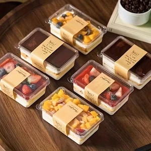 May include: An assortment of individual desserts in clear plastic containers, each with a different topping. Some have fruit like strawberries, blueberries, and mangoes. Others have chocolate. Each container is wrapped with a brown paper band with text.