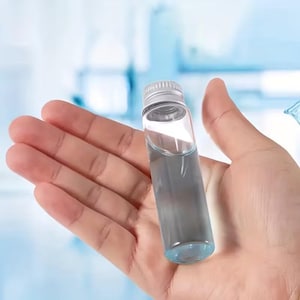 May include: A small, clear glass vial with a silver cap, filled with a blue-tinted liquid. The vial is held in an open hand against a soft blue background. The vial appears to be about 7.6 cm in height.
