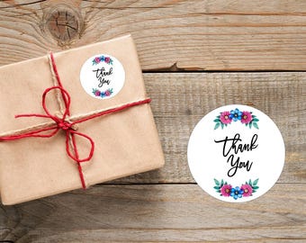 Könnte beinhalten: Ein Geschenk, verpackt in braunes Papier, mit roter Schnur und einem weißen Aufkleber mit der Aufschrift "Thank You". Der Aufkleber ist von einem floralen Design in Rosa, Blau und Grün umgeben. Der Hintergrund ist aus Holz.