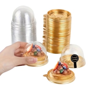 May include: Clear plastic dome-shaped containers with gold-coloured bases, stacked. One container holds a small pastry topped with strawberries and blueberries. A hand holds one of the containers.