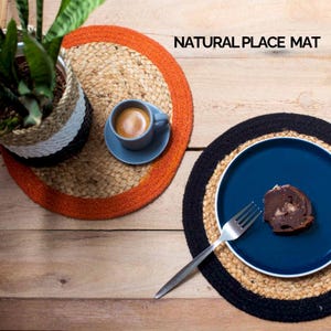 May include: Two round natural jute placemats with contrasting trim. One placemat has an orange trim and the other has a black trim. A blue plate with a chocolate cake slice and a fork is on the black trimmed placemat. A cup of coffee is on the orange trimmed placemat.