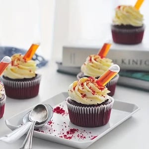 May include: Several red velvet cupcakes with white frosting and a small glass tube filled with orange liquid. The cupcakes are on a white tray with spoons and red sprinkles. The cupcakes are in a white paper cup.