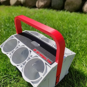 Könnte beinhalten: Ein grauer, 3D-gedruckter Motorblock mit sechs kreisförmigen Öffnungen, einer schwarzen Oberseite und einem roten Griff. Der Griff ist gebogen und an den Seiten befestigt. Der Text "3D Idejos Tau" ist auf der schwarzen Oberseite aufgedruckt.