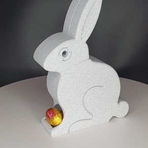 以下が含まれることがあります： 白いセラミック製のイースターバニーで、中に金色のチョコレートの卵が入っています。バニーは白い表面に座っています。