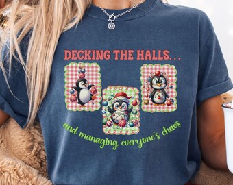 May include: A blue t-shirt with the text "Decking the Halls... and managing everyone's chaos." The shirt features three penguin illustrations with Christmas decorations. The penguins are in decorative frames with red and green accents.