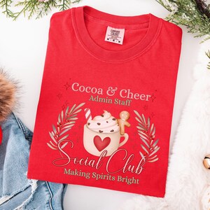 May include: A red t-shirt with the text "Cocoa & Cheer Admin Staff" and "Social Club Making Spirits Bright." The graphic features a mug of hot cocoa with whipped cream, a candy cane, and a gingerbread man. The shirt is folded.