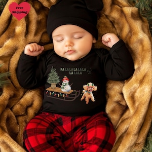 May include: A baby wearing a black long-sleeved shirt and red plaid pants. The shirt has a Christmas tree, snowman, and dog graphic with the text "FA LA LA LA LA LA LA LA LA." The baby is also wearing a black hat.