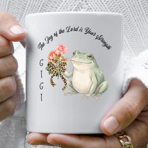 May include: White ceramic mug with a green frog holding a bouquet of pink and orange flowers and a leopard print bow. The mug has the text "The Joy of the Lord is Your Strength" and the name "GIGI".