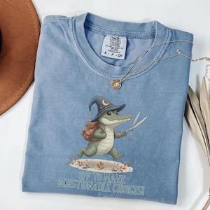 May include: A light blue Comfort Colors t-shirt featuring a cartoon alligator wearing a witch hat and backpack, holding scissors. The text "OFF TO MAKE QUESTIONABLE CHOICES!" is printed below the alligator. A gold necklace is draped across the shirt.