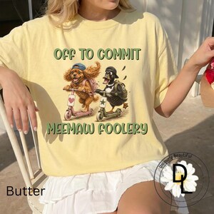 May include: A pale yellow t-shirt featuring a graphic of a brown spaniel and a black duck riding scooters. The dog wears a blue cap and pink backpack, while the duck wears a black cap and green backpack. Text reads: OFF TO COMMIT MEEMAW FOOLERY.