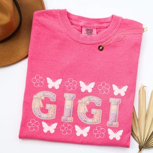 May include: A pink Comfort Colors t-shirt with the name "GIGI" in a patchwork design, adorned with white butterflies and flowers. A gold necklace with a pendant rests on the shirt. A brown hat is visible in the upper left corner.