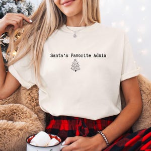 May include: A cream-coloured t-shirt featuring the text "Santa's Favorite Admin" and a small black Christmas tree design. The shirt is made of a soft material, perfect for the festive season.