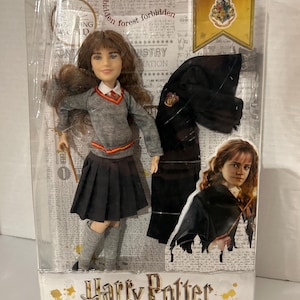 May include: A Hermione Granger doll from the Harry Potter series, packaged in a clear plastic box. The doll wears a gray sweater, black skirt, and gray knee socks. The packaging includes the text "Harry Potter" and "Hermione Granger".