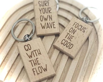 Könnte beinhalten: Drei Holzschlüsselanhänger mit inspirierenden Sprüchen, die darauf graviert sind. Die Schlüsselanhänger sind "Surf Your Own Wave", "Focus On The Good" und "Go With The Flow".