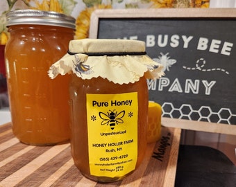 Puede incluir: Dos tarros de vidrio de miel, uno con una etiqueta amarilla y negra que dice "Pure Honey" y "Unpasteurized Honey Holler Farm Rush, NY (585) 439-4729 Honeyhollerform@yahoo.com 680 g Net Weight 24 oz", y el otro con un paño blanco que cubre la tapa. Los tarros están sobre una superficie de madera con una pizarra negra detrás que dice "The Busy Bee Company".