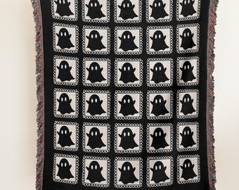 May include: Black woven blanket featuring a repeating pattern of white-framed ghost illustrations. The blanket has a fringed edge in shades of black, white, and red. The blanket is held up against a white wall and brown floor.