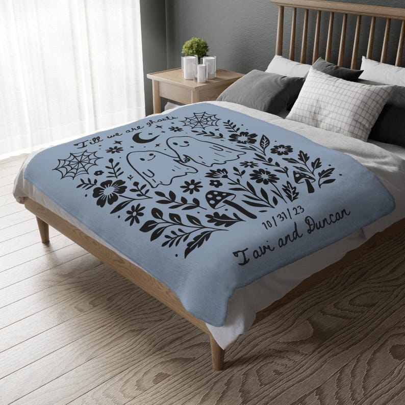 May include: A light blue blanket featuring a black graphic design of two ghosts, flowers, mushrooms, and spiderwebs. The text "Till we are ghosts" and "10/31/23 I avi and Duncan" are also included in the design. The blanket is on a bed.