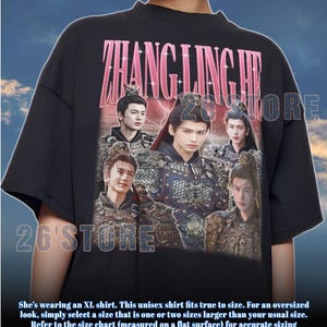 May include: Black unisex t-shirt featuring a graphic of several people in ornate costumes with the text "ZHANG LING HE" in pink. The shirt also has the text "26'STORE".