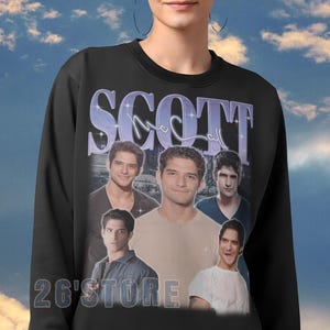 K&ouml;nnte beinhalten: Schwarzes Sweatshirt mit dem Namen "SCOTT" in einer violetten, stilisierten Schriftart. Das Sweatshirt zeigt mehrere Bilder einer Person. Der Text "26'STORE" ist unten aufgedruckt. Der Hintergrund ist ein blauer Himmel mit Wolken.
