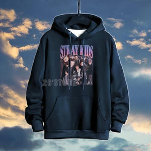 May include: Navy blue hoodie featuring the text "STRAY KIDS" in a purple font above a group photo. The hoodie has a front pocket and the text "26'Store" at the bottom. The background is a cloudy sky.