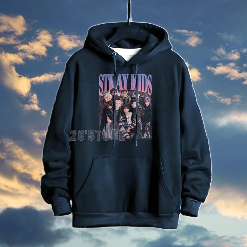 May include: Navy blue hoodie featuring the text "STRAY KIDS" in a purple font above a group photo. The hoodie has a front pocket and the text "26'Store" at the bottom. The background is a cloudy sky.