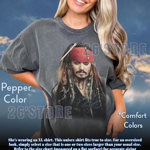 May include: Pepper-colored unisex t-shirt featuring a graphic of a character with a red bandana and dreadlocks. The shirt has the text "26'STORE" and "Comfort Colors". The shirt is oversized and fits true to size.