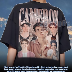 May include: Black t-shirt featuring a collage of images of Cameron Boyce, with the name "CAMERON" in a metallic font across the top. The shirt includes the text "Boyce" in a cursive font. The shirt is unisex.