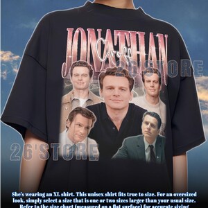 Pode incluir: Camiseta unissex preta com uma colagem de imagens e o nome "Jonathan Groff" em rosa e branco. A camiseta tem o texto "26'STORE" impresso. A camiseta é tamanho XL.