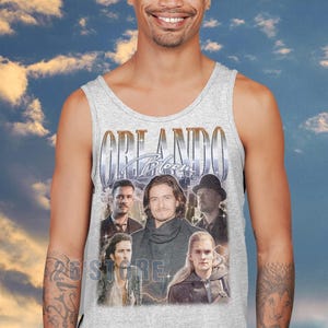 May include: A light gray tank top featuring a collage of images of Orlando Bloom with the text "ORLANDO Bloom" in a gold font. The tank top is worn by a person smiling in front of a cloudy sky background.