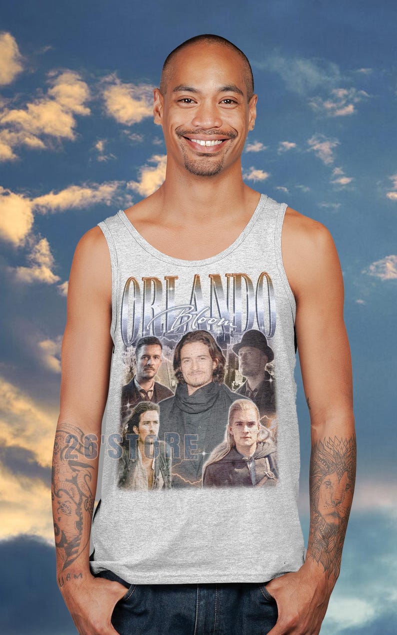 May include: A light gray tank top featuring a collage of images of Orlando Bloom with the text "ORLANDO Bloom" in a gold font. The tank top is worn by a person smiling in front of a cloudy sky background.