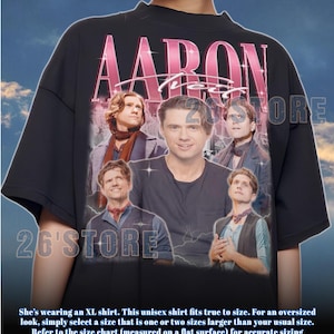 May include: Black unisex t-shirt featuring a collage of images and the name "AARON" in pink. The shirt includes the text "26'STORE" and a sizing guide. The shirt is an XL size.