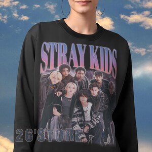 May include: Black sweatshirt featuring a group of people with the text "STRAY KIDS" in a gradient font. Below the text is the text "26'STORE" in a white font. The sweatshirt has a crew neck and long sleeves.