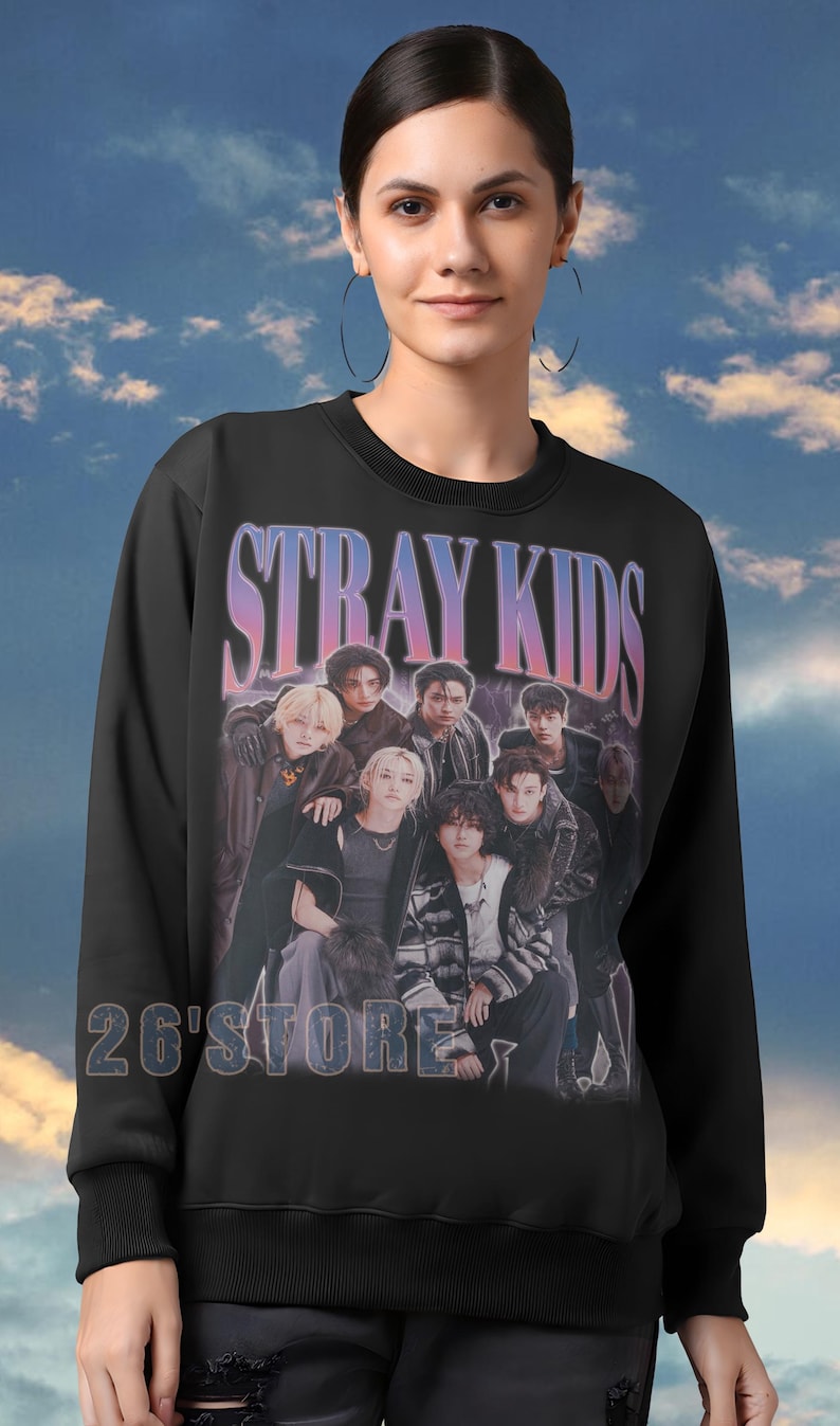 May include: Black sweatshirt featuring a group of people with the text "STRAY KIDS" in a gradient font. Below the text is the text "26'STORE" in a white font. The sweatshirt has a crew neck and long sleeves.