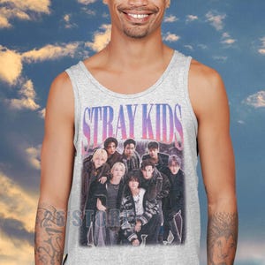 May include: A light gray tank top featuring a group of people with the text "STRAY KIDS" in a purple and pink gradient. The tank top is worn by a person with a blue sky background.